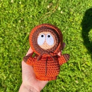 Puede incluir: Muñeco de ganchillo hecho a mano de un personaje de dibujos animados, con una chaqueta con capucha naranja, detalles marrones y ojos blancos grandes. El muñeco se sostiene sobre hierba verde, mostrando la artesanía detallada.