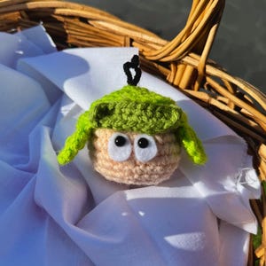 May include: A crocheted doll with a light tan body, large white eyes, and a green hat. The doll has a black stem and is resting on a white cloth inside a wicker basket.