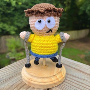 Puede incluir: Una figura de ganchillo con cabello castaño, grandes ojos de fieltro blanco y una pequeña boca negra. La figura viste una camisa amarilla brillante y pantalones azules, y sostiene dos muletas grises de ganchillo sobre una base de madera.