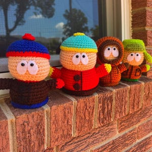 May include: Four handmade crochet dolls, each representing a character from a popular animated series. The dolls are displayed on a brick ledge. Each doll has a unique outfit and hat, with large, white eyes and small arms.