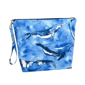 May include: A blue zippered pouch with a wrist strap, featuring a pattern of white and navy blue whales against a watercolor-style blue background. The pouch is rectangular with a navy blue zipper and strap.