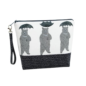 May include: A white zippered pouch featuring a repeating black and white illustration of three bears, each holding an open umbrella. The bottom section of the pouch is black with a white dotted pattern, and it includes a matching black wrist strap.