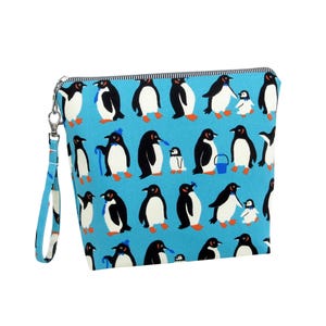 May include: A blue zippered pouch with a repeating pattern of cartoon penguins in various poses. The penguins are black and white with orange feet and beaks. The pouch has a blue wrist strap and a black and white striped zipper.