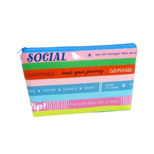 May include: A colorful canvas zippered pouch with horizontal stripes in pink, green, blue, and yellow. The pouch features text including "SOCIAL", "trust your journey", and "MADE IN JAPAN". A white bird graphic is also present.