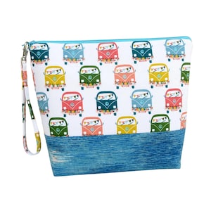 May include: A white zippered pouch with a blue denim-look bottom and a wrist strap. The pouch is patterned with colorful vintage camper vans. Each van has a peace sign and a banner of colorful flags.