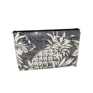 May include: A black and white zippered pouch featuring a pineapple and leaf pattern. The pouch has a black zipper and is made of a textured fabric. The design is a tropical theme, suitable for cosmetics or small items.