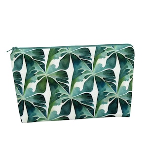 May include: A rectangular cosmetic bag with a watercolor-style pattern of large green leaves on a white background. The bag has a teal zipper and trim. The design features a repeating pattern of tropical leaves.