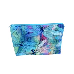 May include: A rectangular cosmetic bag with a blue zipper. The bag features a watercolor design of blue dragonflies with detailed wings against a colorful background of green, pink, and purple.