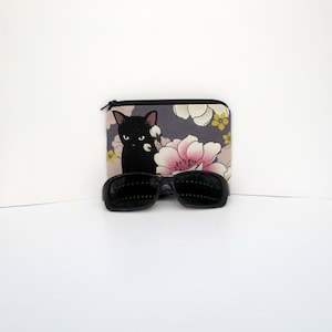 May include: A black cat with yellow eyes is printed on a gray fabric pouch with pink and yellow flowers. The pouch is closed with a zipper. A pair of black sunglasses are in front of the pouch.