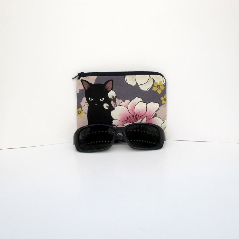 May include: A black cat with yellow eyes is printed on a gray fabric pouch with pink and yellow flowers. The pouch is closed with a zipper. A pair of black sunglasses are in front of the pouch.