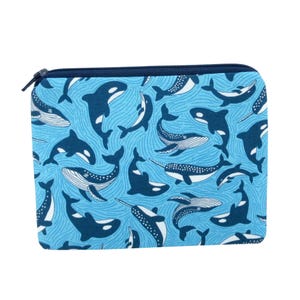 May include: A blue zippered pouch featuring a pattern of various whale illustrations in white and navy blue. The whales are set against a light blue background with wavy lines. The zipper is navy blue.