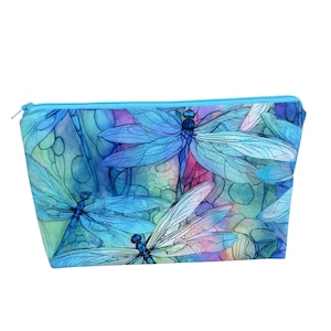 May include: A rectangular cosmetic bag with a blue zipper. The bag features a watercolor design of blue dragonflies with detailed wings against a backdrop of blue, purple, and green hues. The bag is likely made of fabric.