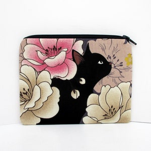 May include: Black cat with white eyes peeking out from behind pink and beige floral print fabric. The fabric has a zipper closure.