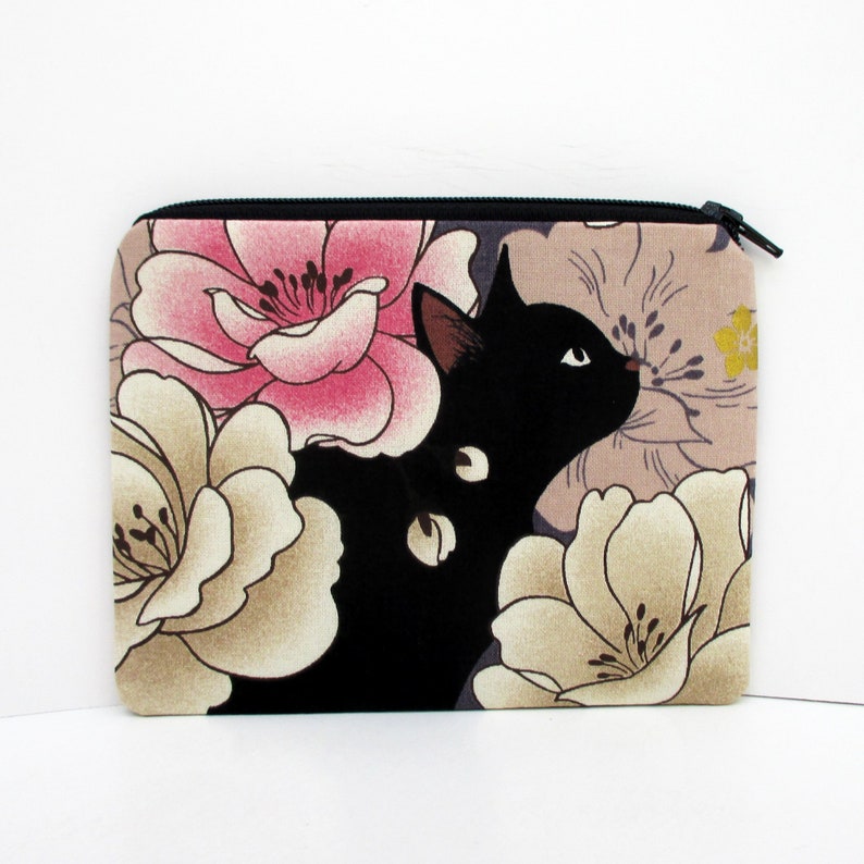 May include: Black cat with white eyes peeking out from behind pink and beige floral print fabric. The fabric has a zipper closure.