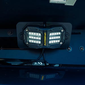 Può includere: Una barra luminosa a LED rettangolare nera con due pannelli luminosi distinti e un logo giallo centrale, montata su una superficie scura. Sopra la luce, un connettore elettrico grigio è collegato a un filo rosso e nero, con riflesso visibile.