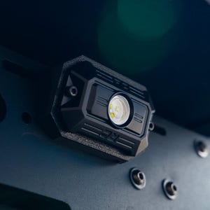 Può includere: Un modulo luce LED rettangolare nero con finitura testurizzata e una lente circolare centrale. L'alloggiamento presenta le lettere RC in rilievo sul bordo inferiore. È montato su una staffa metallica nera fissata con bulloni a vista.
