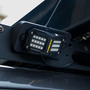 Puede incluir: Barra de luces LED negra con el texto "AUXBEAM" en amarillo. La barra de luces está montada en un soporte de metal negro. La barra de luces tiene múltiples luces cuadradas pequeñas.