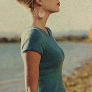 May include: A vintage-style image of a woman wearing a teal knit top and dark purple pants. She has blonde hair and is wearing pink earrings. The background shows a body of water and a shoreline.
