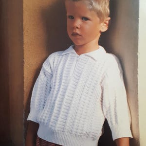 Puede incluir: Un niño pequeño con un suéter de punto blanco con cremallera y una falda de cuadros rojos y blancos.