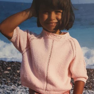 May include: A young girl with dark hair and a pink sweater smiles at the camera. She is standing on a beach with a rocky shore in the background.