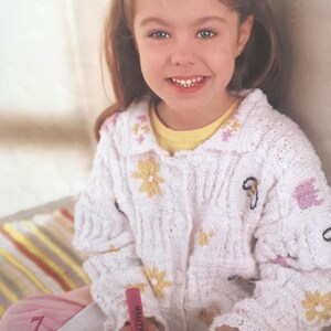 May include: A young person wearing a white knitted cardigan decorated with yellow and pink floral patterns. The cardigan features a collared neckline and button fastenings. The person is holding a pink highlighter and writing on a notepad. The number 7 is visible.