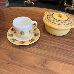 以下が含まれることがあります： 緑の葉のデザインが施された黄色の陶器製のティーカップとソーサーのセット。セットは木製のテーブルの上に置かれています。