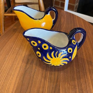 Könnte beinhalten: Zwei Keramik-Saucieren mit blauen und gelben Designs. Eine Sauciere hat ein Sonnen- und Palmenmotiv, die andere hat eine einfache gelbe Glasur.