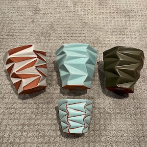 May include: Four ceramic planters in different colours. The planters are all the same shape, with a geometric design. The colours are white and brown, light blue, and dark green.