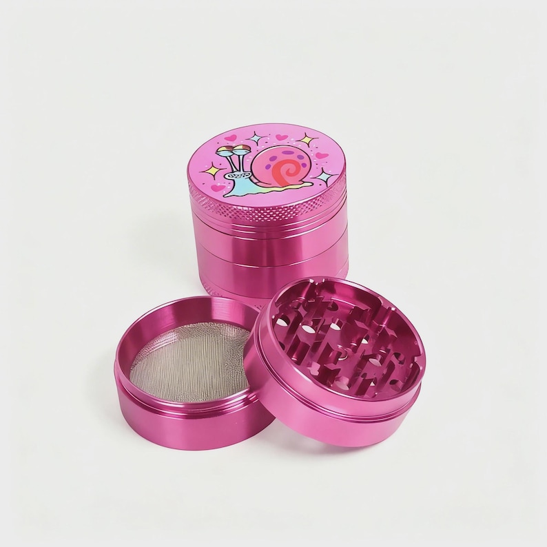 May include: A disassembled pink metal grinder with a cartoon snail design on the top. The grinder has multiple parts, including a mesh screen and a section with sharp teeth for grinding.