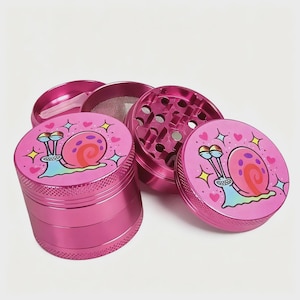 May include: A disassembled pink metal grinder with a cartoon snail design. The grinder is in several pieces, including a top, bottom, and a mesh screen. The snail has a pink shell and is surrounded by hearts and stars.
