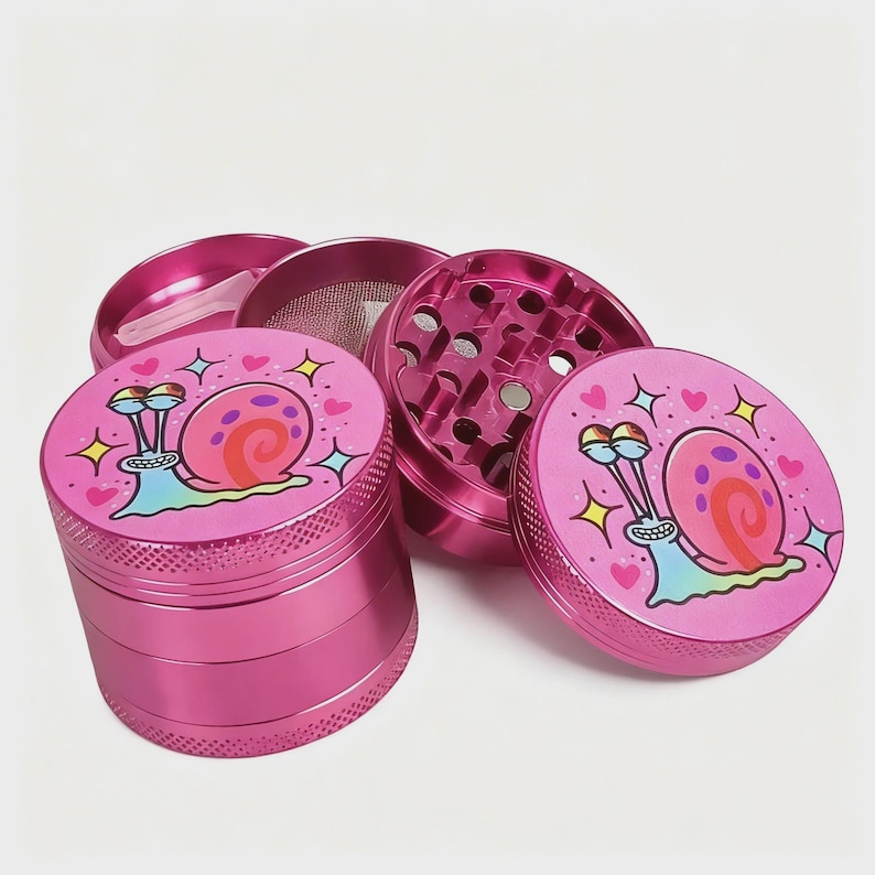 May include: A disassembled pink metal grinder with a cartoon snail design. The grinder is in several pieces, including a top, bottom, and a mesh screen. The snail has a pink shell and is surrounded by hearts and stars.