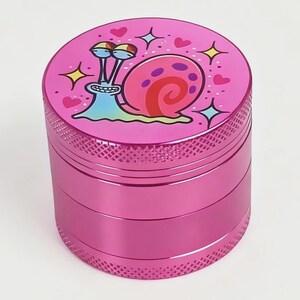 May include: A pink, three-piece grinder with a cartoon snail design on the top. The snail has a pink shell, blue body, and is surrounded by hearts and stars. The grinder has a textured, ridged exterior.