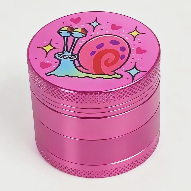 May include: A pink, three-piece grinder with a cartoon snail design on the top. The snail has a pink shell, blue body, and is surrounded by hearts and stars. The grinder has a textured, ridged exterior.