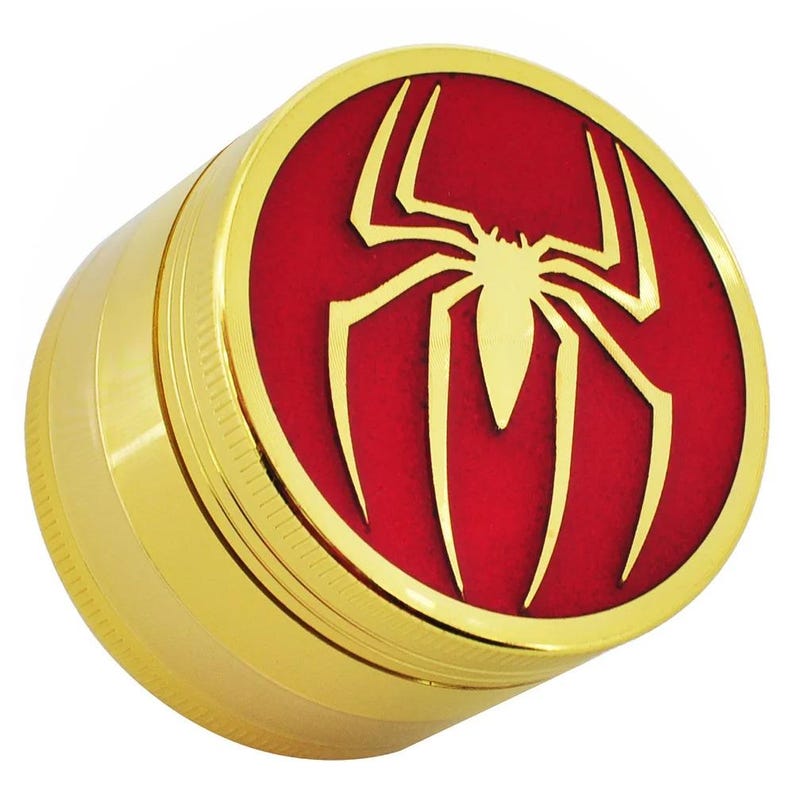 May include: A gold-colored, cylindrical grinder with a red top featuring a gold spider emblem. The grinder has a ribbed texture on the side and a shiny, metallic finish. The spider emblem is a prominent design element.