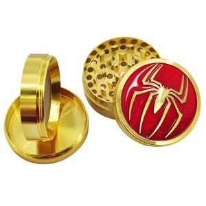 May include: A gold-colored, three-piece herb grinder. The top piece features a red emblem with a gold spider design. The grinder is disassembled, showing the various components, including a mesh screen and a grinding chamber.