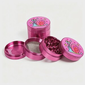 May include: A disassembled pink metal grinder with four separate pieces. The top piece features a cartoon snail design with hearts and stars. The grinder has a mesh screen and a grinding chamber with diamond-shaped teeth.