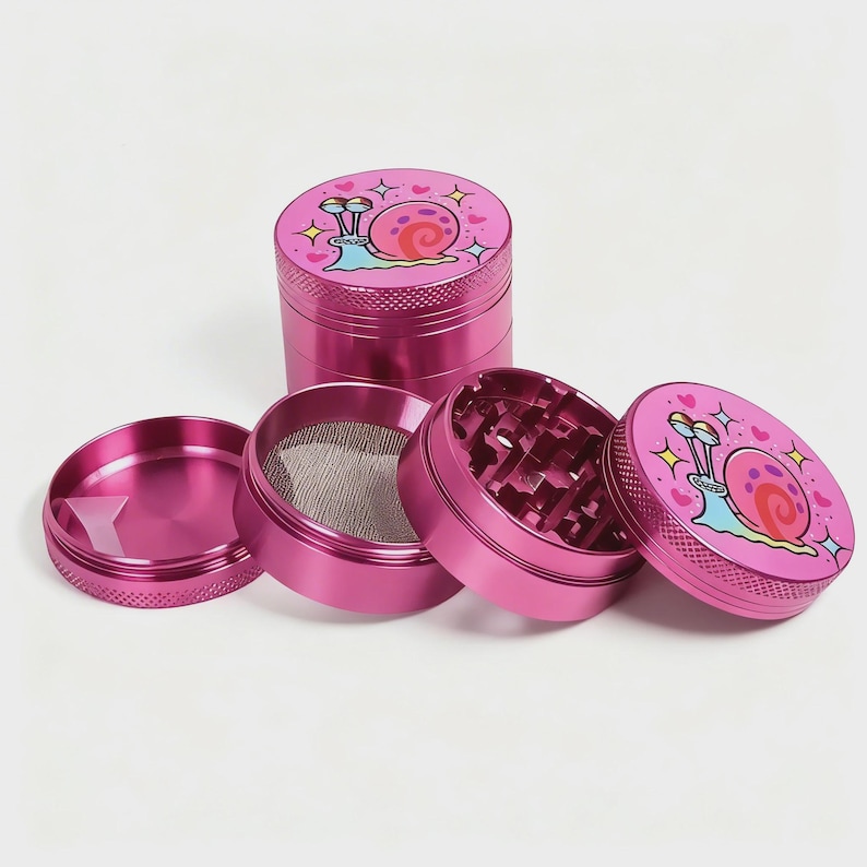 May include: A disassembled pink metal grinder with four separate pieces. The top piece features a cartoon snail design with hearts and stars. The grinder has a mesh screen and a grinding chamber with diamond-shaped teeth.