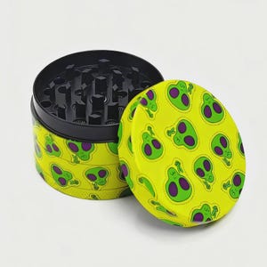 May include: A round, two-piece grinder with a black interior and a bright yellow exterior. The exterior is covered in a repeating pattern of green alien heads with purple eyes. The lid is slightly ajar, revealing the grinding teeth.