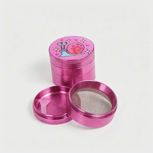 May include: A disassembled pink metal grinder with a cartoon snail design on the top. The grinder is in three parts, with a mesh screen and a small plastic scraper visible. The snail has a pink shell and is surrounded by hearts.