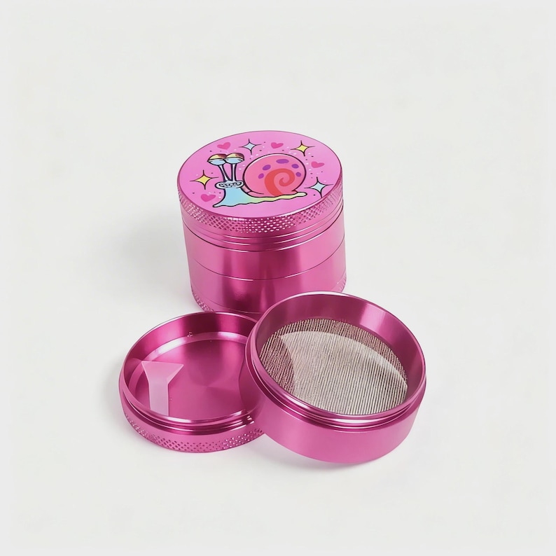 May include: A disassembled pink metal grinder with a cartoon snail design on the top. The grinder is in three parts, with a mesh screen and a small plastic scraper visible. The snail has a pink shell and is surrounded by hearts.