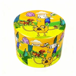 Peut inclure: Un broyeur cylindrique jaune orné d'un motif de paysage de dessins animés. Le design comprend un personnage jaune ressemblant à un chien avec de longs bras, un garçon en chemise bleue et d'autres éléments colorés. Le broyeur est sur fond blanc.