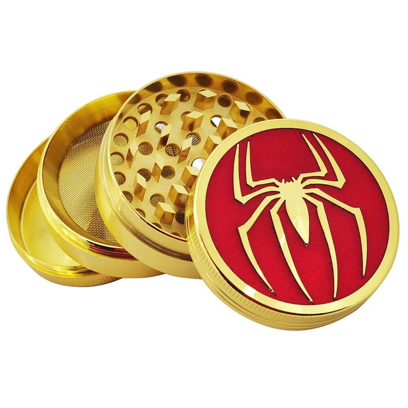 May include: A gold-colored, four-piece herb grinder. The top piece features a red circle with a gold spider emblem. The grinder is open, revealing the mesh screen and grinding teeth. The design is a nod to the Spider-Man character.