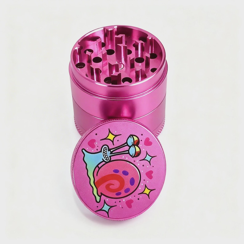 May include: A pink metal grinder with a lid featuring a cartoon snail character. The grinder is cylindrical with a ridged base and top. The lid has a colorful illustration of a snail with hearts and stars.
