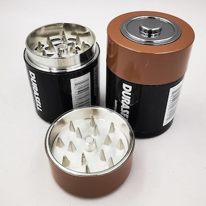 Peut inclure: Un moulin à herbes cylindrique conçu pour ressembler à une pile de type D avec un extérieur noir et cuivre. L'appareil présente un intérieur en argent métallique avec des dents pointues en forme de pyramide. Le côté affiche le texte DURASELL.