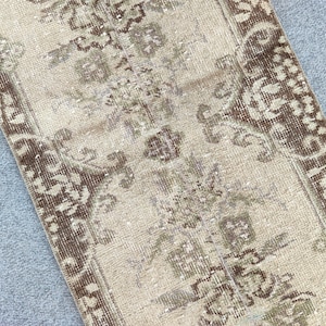 May include: A rectangular, vintage-style rug with a distressed appearance. The rug features a central field of beige with intricate floral and geometric patterns in shades of brown and olive green. The edges are bordered with a darker brown.