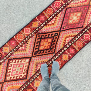 May include: A vibrant, rectangular runner rug with a geometric pattern in shades of red, orange, and black. The rug features diamond and square motifs, creating a bold and colorful design. The rug is placed on a gray surface.