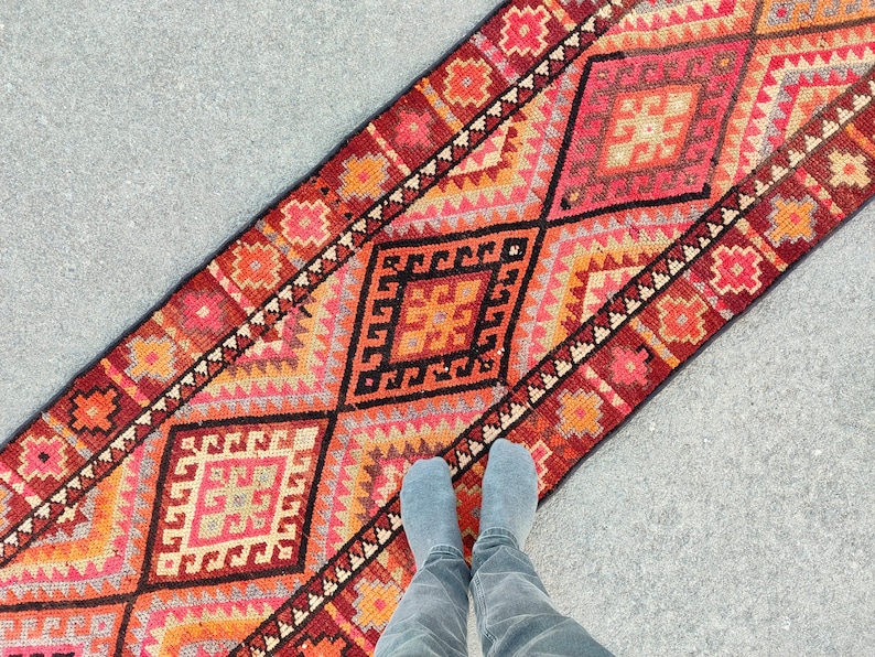 May include: A vibrant, rectangular runner rug with a geometric pattern in shades of red, orange, and black. The rug features diamond and square motifs, creating a bold and colorful design. The rug is placed on a gray surface.