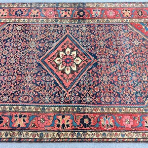 Peut inclure: Un tapis rectangulaire pr&eacute;sentant un champ central bleu fonc&eacute; avec un grand m&eacute;daillon en forme de losange contenant un motif floral en cr&egrave;me, rouge et bleu. Le tapis est encadr&eacute; par une large bordure rouge aux motifs floraux et g&eacute;om&eacute;triques.