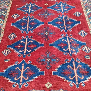 Op de afbeelding: Een rood vloerkleed met een herhalend geometrisch patroon in blauw en crème. Het vloerkleed heeft ruitvormen en een rand met ingewikkelde ontwerpen. De textuur van het vloerkleed is zichtbaar, wat wijst op een geweven constructie.
