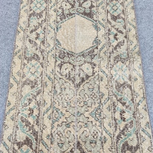 Könnte beinhalten: Ein rechteckiger Teppich im Vintage-Stil mit einem zentralen Rautenmuster. Der Teppich hat einen beigefarbenen Hintergrund mit komplizierten Mustern in Braun und Türkis. Die Ränder haben ein verblasstes Aussehen, was ihm einen Used-Look verleiht.